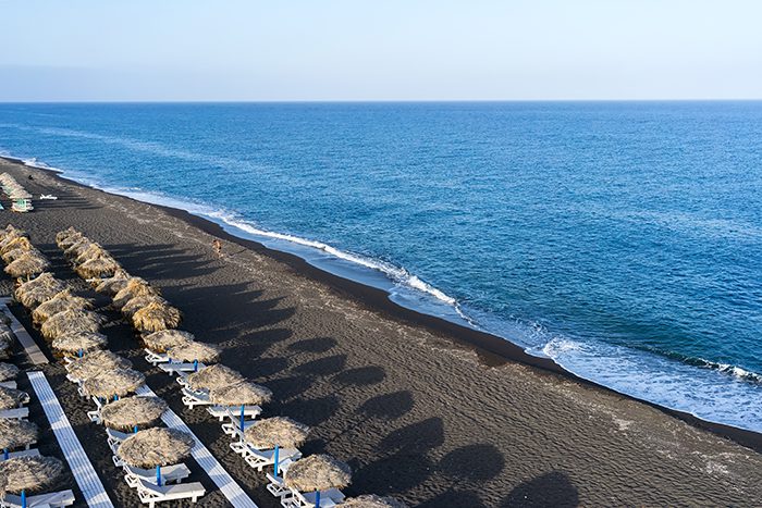 Top view of Perissa beach on the Greek island of Santorini with sunbeds and umbrellas. Beach is covered with fine black sand, and drops off sharply into the water.